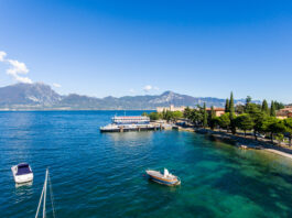 Torri del Benaco festeggia i 50 anni di Federalberghi Garda Veneto Scena di paesaggio con acqua, barche e montagne in background sotto un cielo limpido.