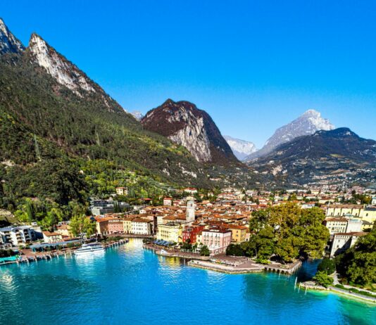 Longenica: a Riva del Garda nasce la fiera sulla longevità Vista panoramica di una città lungo un lago, con montagne verdi e cieli limpidi.