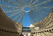 Progetto Altogarda: installate le ultime tele nel biotopo del Muse, mostra in primavera a Riva del Garda Interno di un edificio con una struttura a griglia che forma una cupola aperta verso il cielo blu.