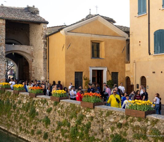Sirmione, 48mila visitatori nel ponte pasquale con il piano Sirmione 2026 Visitatori a Sirmione lungo le mura storiche con fioriere di tulipani.