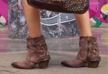 Tendenze calzature 2026: modelli e stili da non perdere Inquadratura in primo piano delle gambe di una persona che indossa stivaletti AS98 in pelle scamosciata marrone con decorazioni di borchie metalliche. La persona tiene una borsa marrone coordinata, posando davanti a un muro con graffiti colorati.