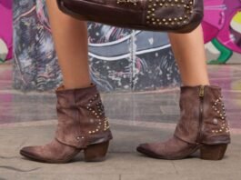 Inquadratura in primo piano delle gambe di una persona che indossa stivaletti AS98 in pelle scamosciata marrone con decorazioni di borchie metalliche. La persona tiene una borsa marrone coordinata, posando davanti a un muro con graffiti colorati.