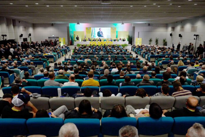 Scena di una grande sala con pubblico seduto davanti a un palco dove un uomo parla
