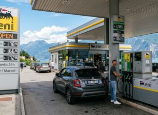 Un distributore di carburante con cartelli sui prezzi, veicoli e una persona in piedi vicino alla pompa.