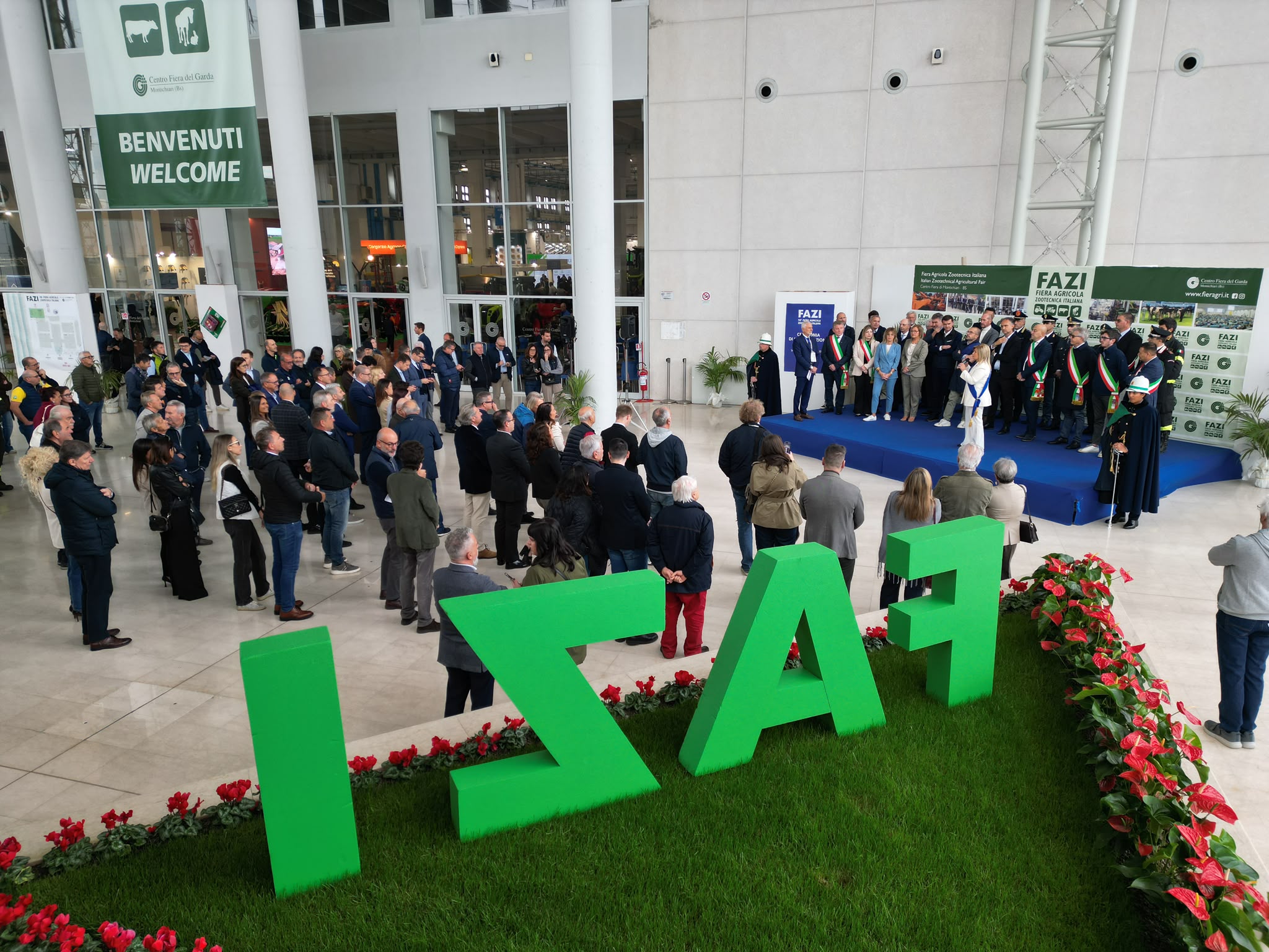 Fazi di Montichiari, inaugurata la 98ª edizione con il ministro Lollobrigida 1 Scena con persone in piedi davanti a un palco e grandi lettere verdi al Centro Fiera di Montichiari per la 98ª Fiera Agricola