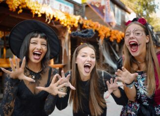 Halloween sul Garda 2026: cosa fare tra Gardaland, Movieland, Sigurtà e le feste in paese Tre persone in abiti a tema festivo sorridono e mostrano le mani in una scena all'aperto con strutture in legno e decorazioni