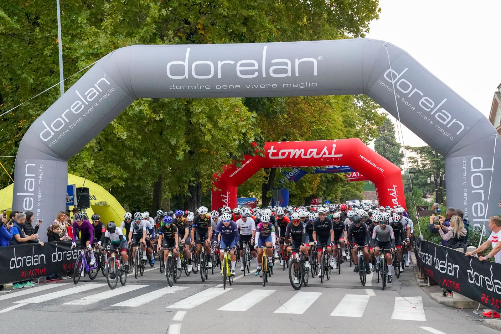 Granfondo Dorelan ReActive a Peschiera del Garda: 1.500 ciclisti da 20 nazioni 1 Corridori in posa davanti a un arco colorato su una strada in un ambiente boscoso.