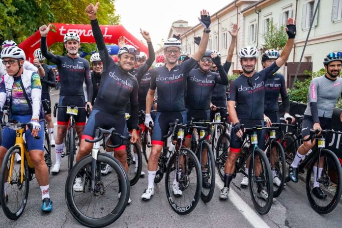 Immagine di Granfondo Dorelan ReActive a Peschiera del Garda: 1.500 ciclisti da 20 nazioni Un gruppo di persone in abbigliamento sportivo con caschi, in piedi su biciclette, con un'insegna rossa alle spalle.