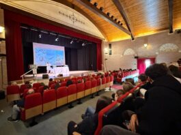 Scena di aula con pubblico seduto e atleti in piedi davanti a schermo per premiazioni a Bardolino