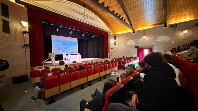 Scena di aula con pubblico seduto e atleti in piedi davanti a schermo per premiazioni a Bardolino