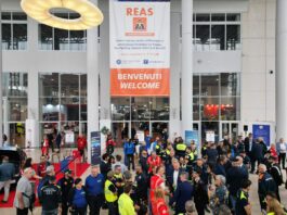 REAS 2026 Montichiari: 310 espositori da 25 Paesi Gruppo di persone davanti a un edificio con una bandiera e una scritta in un ambiente interno.