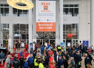 REAS 2026 Montichiari: 310 espositori da 25 Paesi Gruppo di persone davanti a un edificio con una bandiera e una scritta in un ambiente interno.