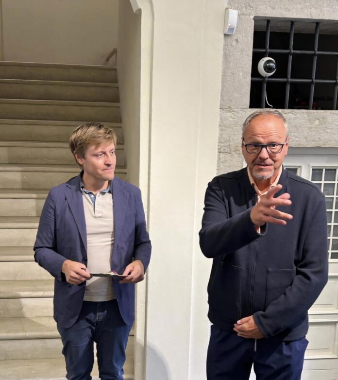 Due persone in abiti diversi stanno in piedi in un ambiente interno con una scala di legno alle loro spalle.