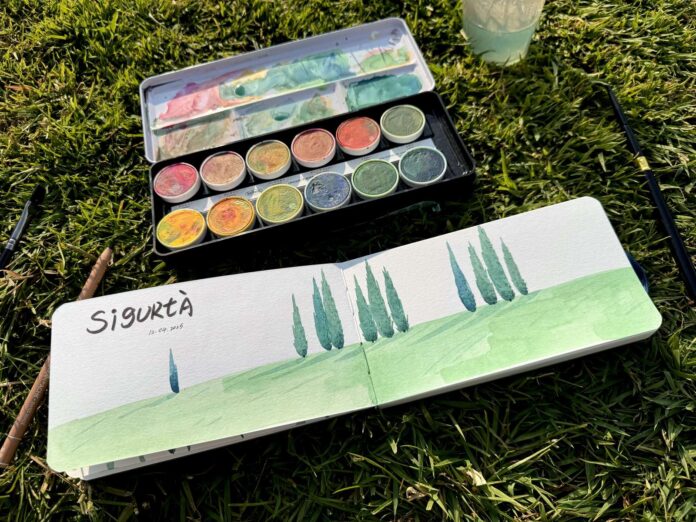 Un taccuino aperto con un disegno di pini su un prato erboso accanto a una scatola di colori